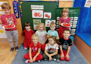 Dzieci siedzą przed tablicą, na której są obrazki przedstawiające pomidory. Dzieci trzymają talerzyki, na których są pokrojone pomidory do degustacji